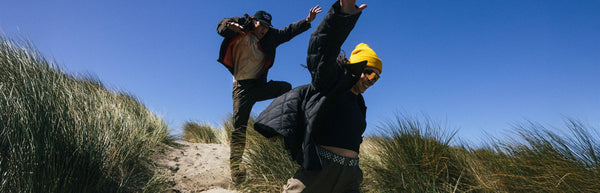 Two people in jackets and hats joyfully leap over grassy sand dunes under a clear blue sky, with tall grass blowing in the wind around them.