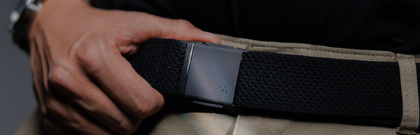 A person’s hand is adjusting a black belt with a metal buckle on beige pants. The person is wearing a black shirt, and part of a wristwatch is visible.