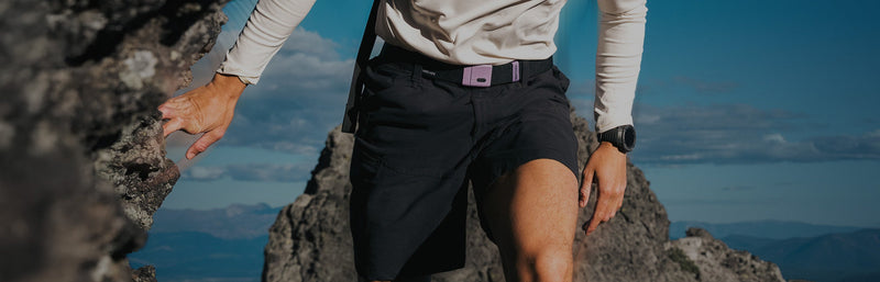A person wearing a long-sleeve shirt, black shorts, and a watch climbs a rocky mountain with blue sky and distant mountains in the background.