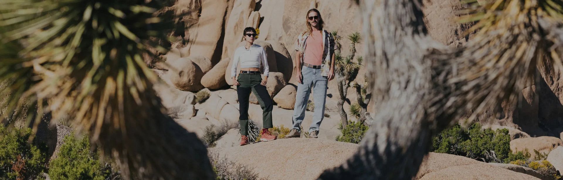 Two people stand on rocks in a desert landscape with large boulders and spiky trees, wearing casual outdoor clothes and sunglasses, surrounded by rugged nature.