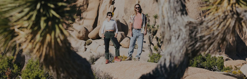 Two people stand on rocks in a desert landscape with large boulders and spiky trees, wearing casual outdoor clothes and sunglasses, surrounded by rugged nature.