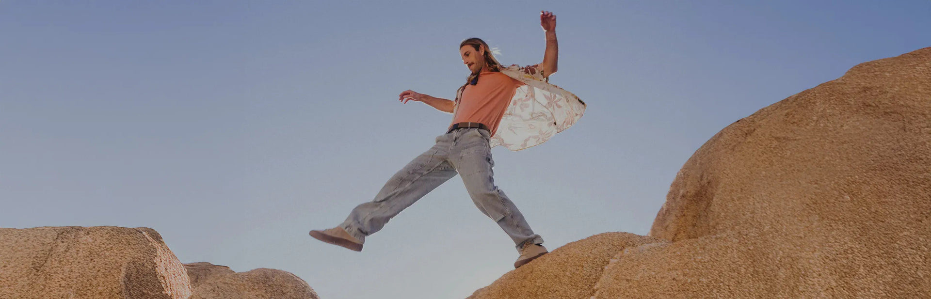 A person with long hair and casual clothes steps with arms outstretched across two large rocks under a clear blue sky.
