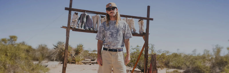 A person wearing a camouflage jacket, a gray shirt, and black pants stands near the ocean with cliffs and blue sky in the background.