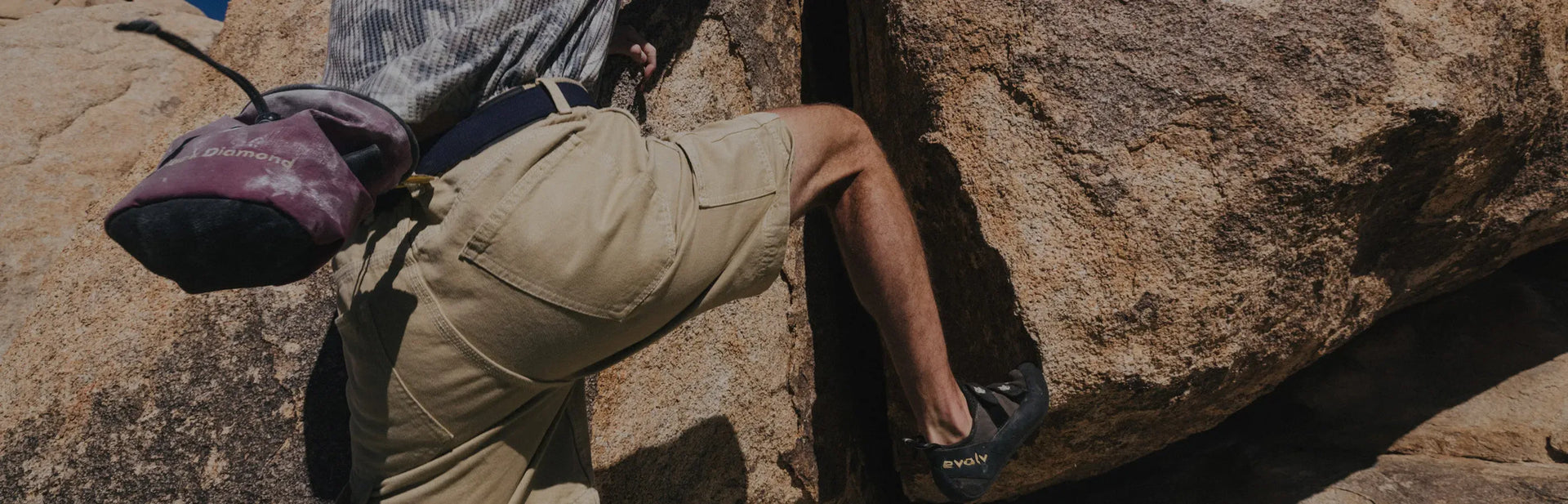 A person stands on a sandy path through tall grass, wearing a black jacket with an orange lining, a white shirt, and olive pants, with blue sky and distant hills in the background.