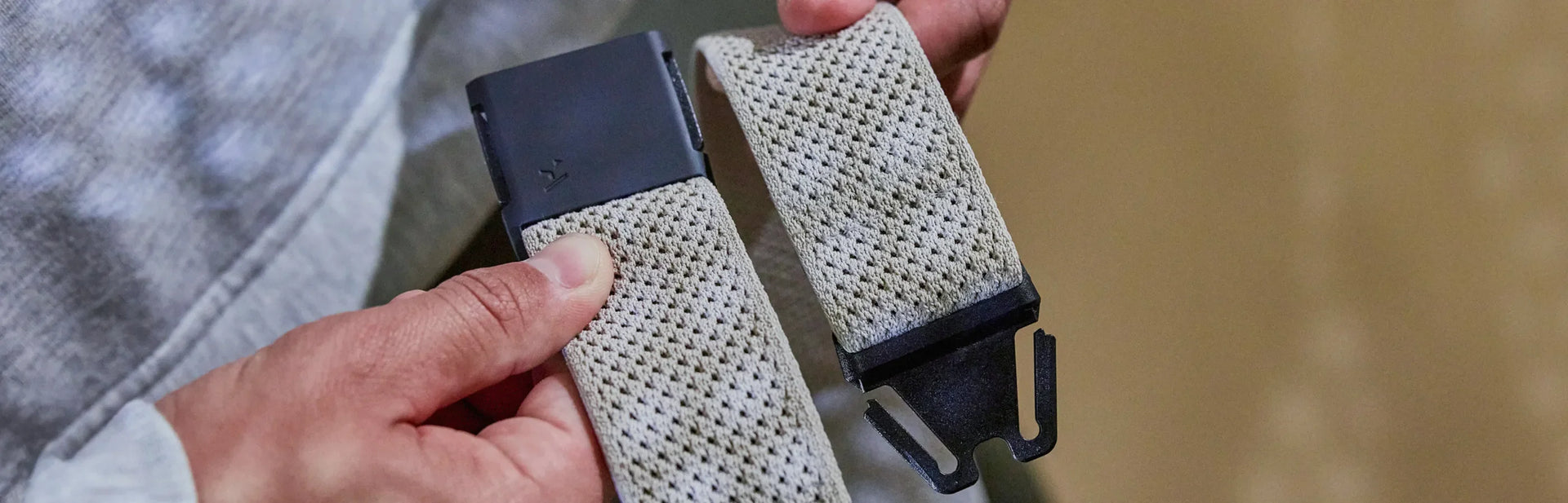 A person’s hand is adjusting a black belt with a metal buckle on beige pants. The person is wearing a black shirt, and part of a wristwatch is visible.