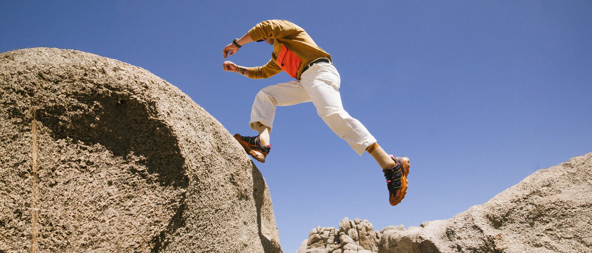 A person in white pants and a brown sweater leaps between large rocky boulders under a clear blue sky.