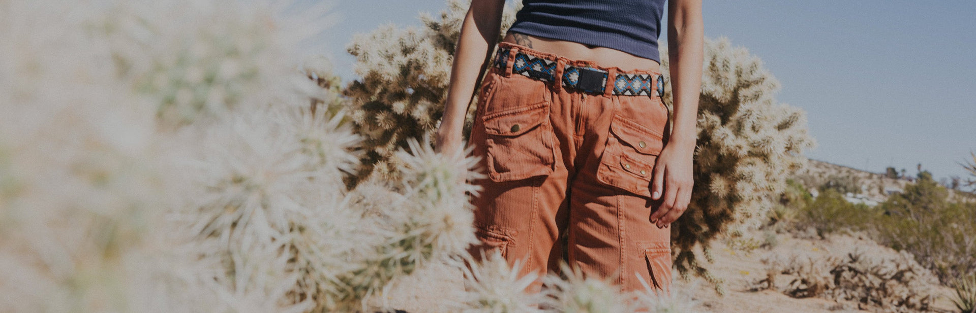 Person wearing orange cargo pants, a blue patterned belt, and a dark tank top standing outdoors near desert plants and cacti under a clear sky.