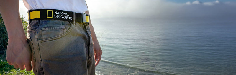A person wearing gray jeans and a National Geographic belt stands by the edge of a grassy cliff, overlooking a calm, hazy ocean under a partly cloudy sky.