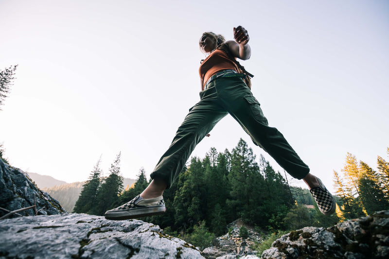 A person in casual clothes steps across rocks outdoors, with tall green trees and a bright sky in the background, photographed from a low angle.
