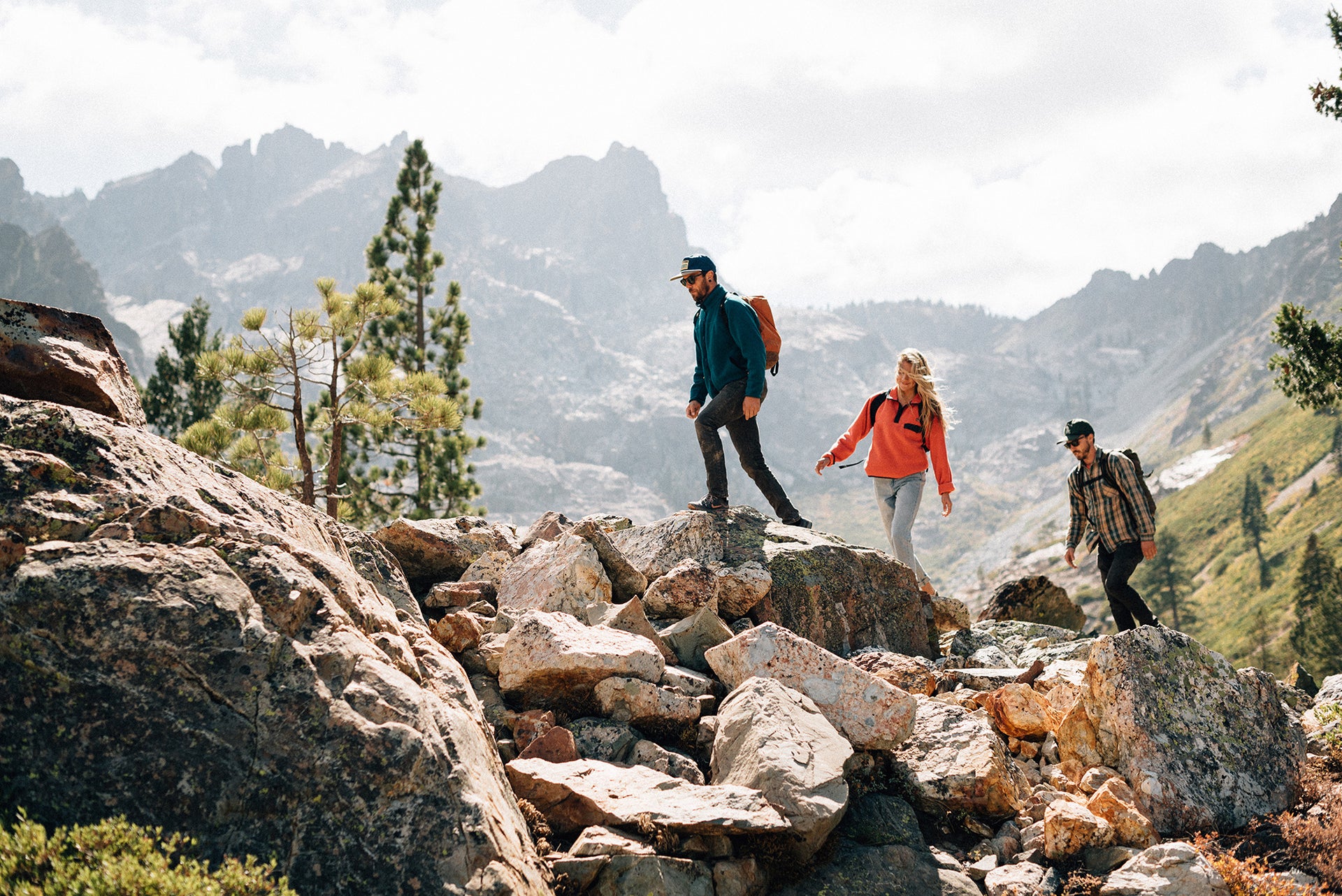 Three people with backpacks hike over rocky terrain in a mountainous landscape, surrounded by pine trees and distant peaks under a partly cloudy sky.