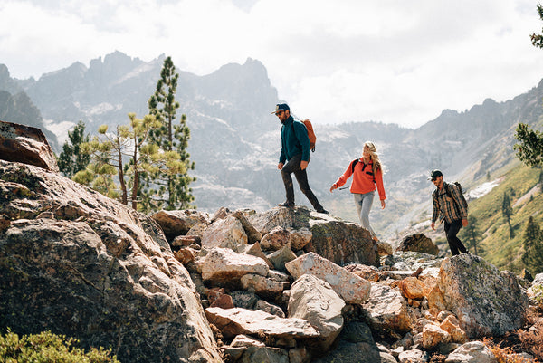 Three people with backpacks hike over rocky terrain in a mountainous landscape, surrounded by pine trees and distant peaks under a partly cloudy sky.