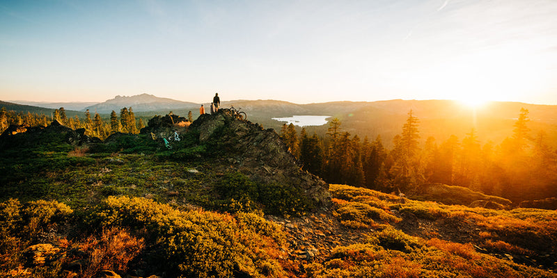 A person stands with a bicycle on a rocky hilltop, overlooking a forest and lake at sunset, with golden sunlight illuminating the landscape and casting long shadows.