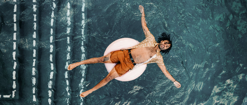 A person in an orange swimsuit and open shirt floats on a pink pool ring in a swimming pool, smiling with arms and legs outstretched.