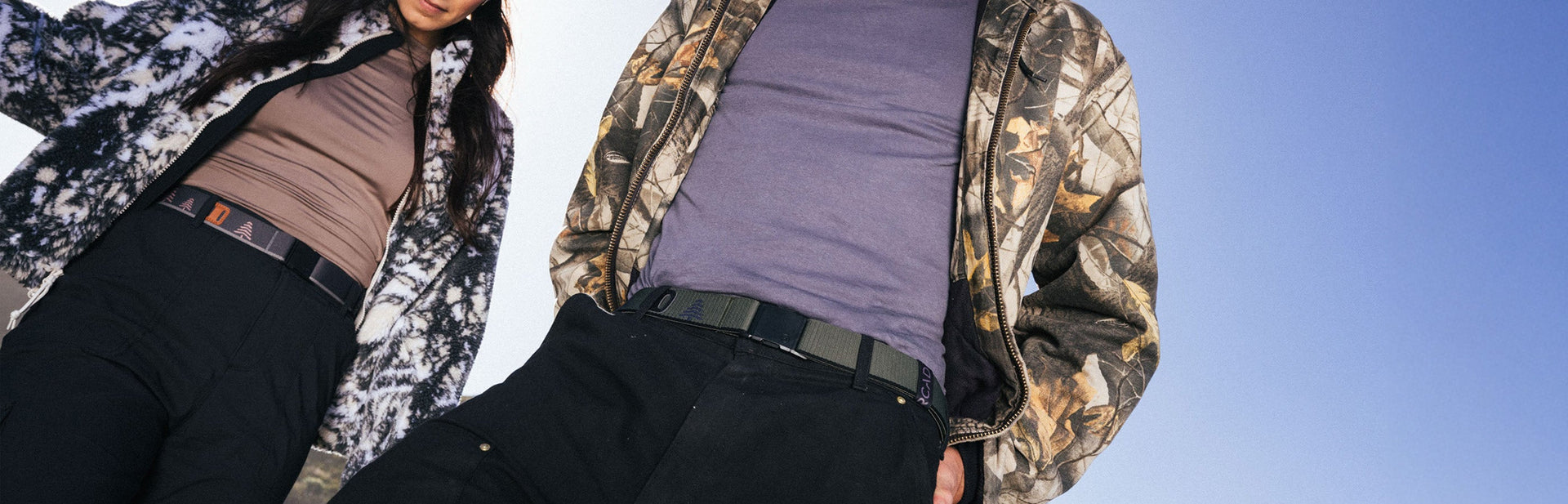 Two people standing outdoors wearing patterned jackets and casual pants, photographed from a low angle against a clear blue sky. Their faces are mostly out of the frame.