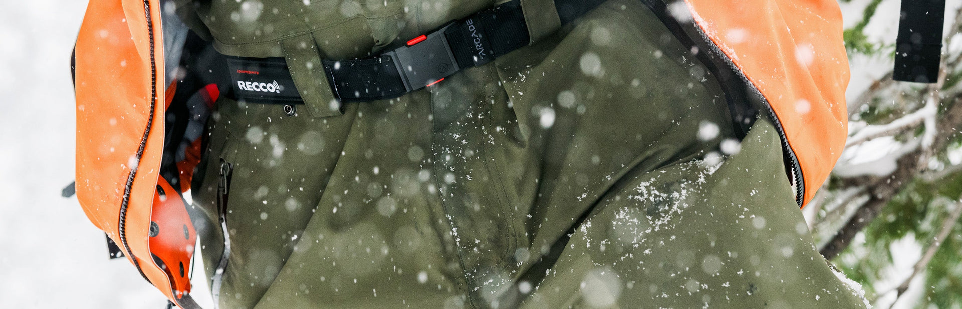 Close-up of a person wearing an orange jacket and green snow pants with a black RECCO belt, standing in falling snow. Snowflakes are visible on the clothing.