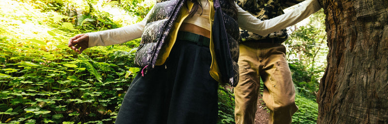 A person wearing tan shorts is caught mid-air while jumping off a rocky cliff, arms and legs extended, against a clear blue sky with hills in the background.