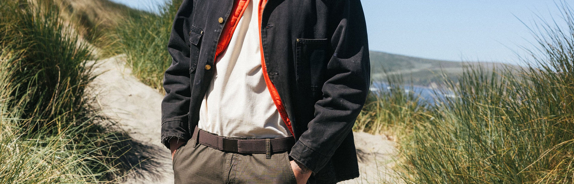 A person stands on a sandy path through tall grass, wearing a black jacket with an orange lining, a white shirt, and olive pants, with blue sky and distant hills in the background.