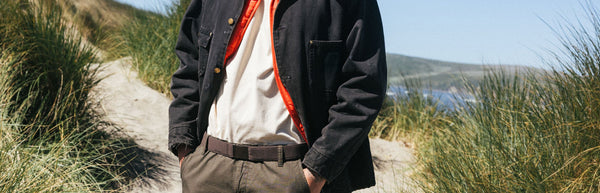 A person stands on a sandy path through tall grass, wearing a black jacket with an orange lining, a white shirt, and olive pants, with blue sky and distant hills in the background.