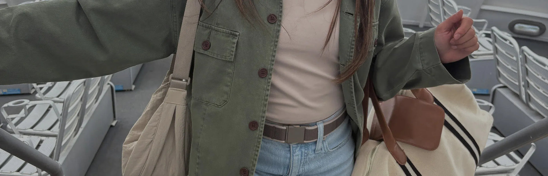 A person in a green jacket and light jeans holds a beige tote bag and a cream duffel bag with brown accents, standing among rows of empty white seats on a bus or train.