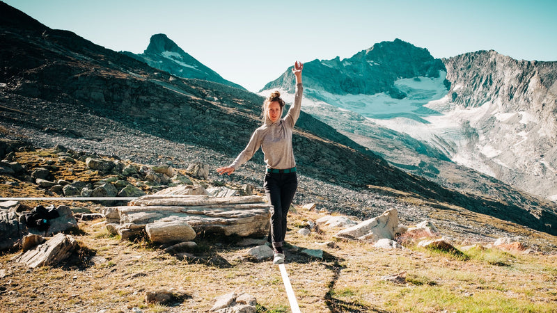 A person balances on a slackline stretched across a grassy mountain landscape with rocky peaks and a glacier in the background under a clear blue sky.