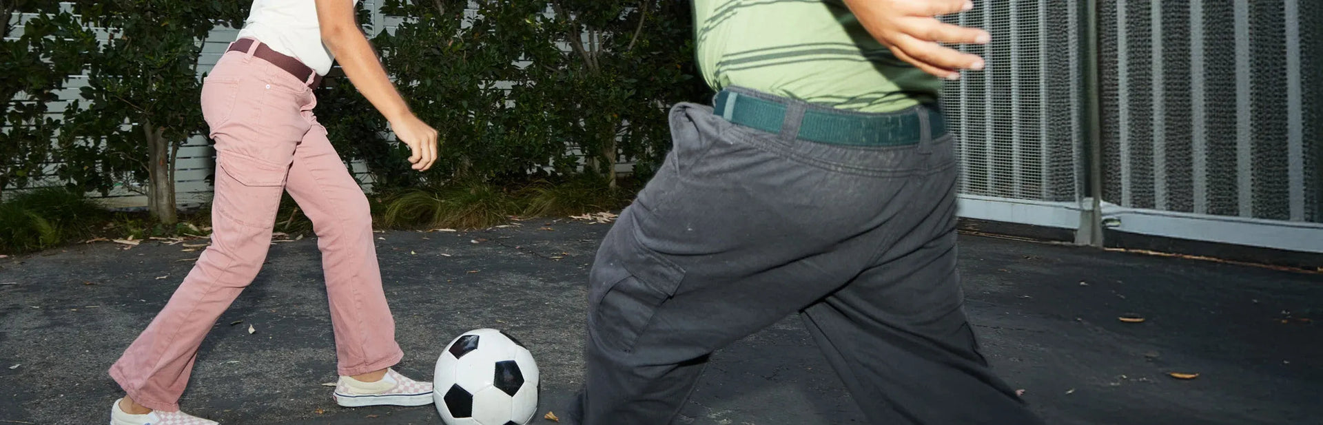 Two people playing soccer outdoors on pavement, one wearing pink pants and sneakers, the other in dark pants and a green shirt, with a black-and-white soccer ball between them.