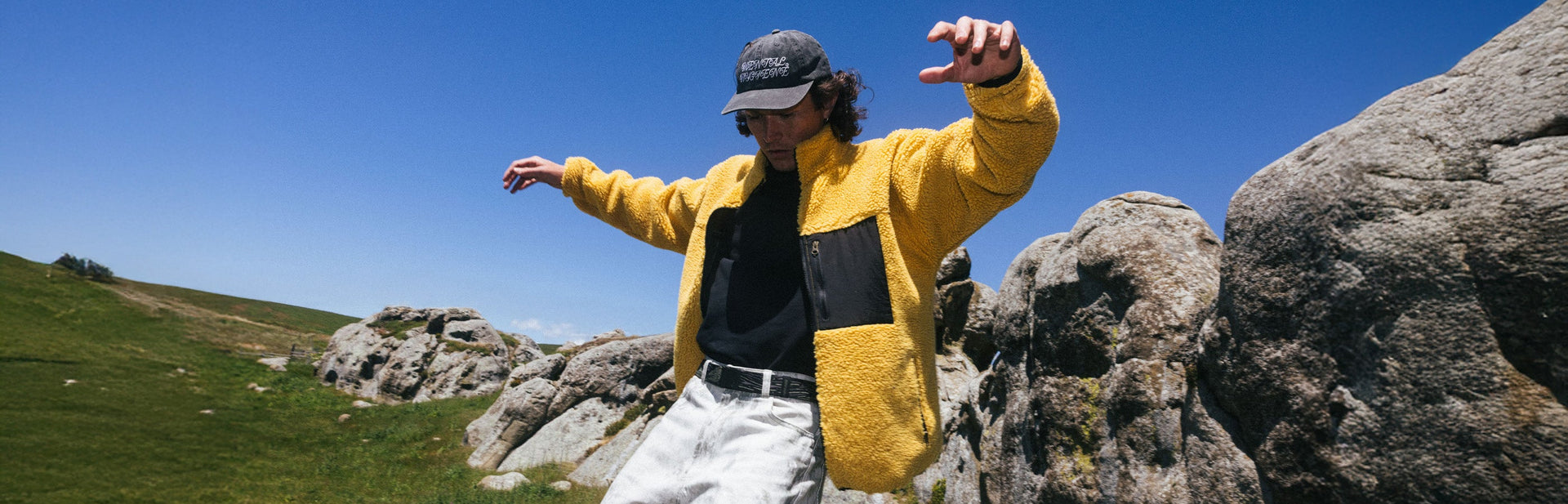 A person in a yellow jacket and black cap balances with arms outstretched on rocks outdoors, with green grass and blue sky in the background.