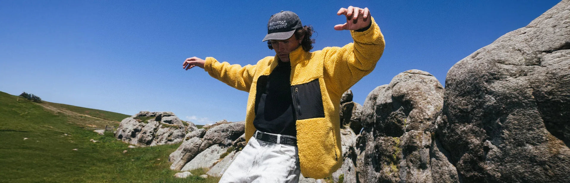 A person in a yellow jacket and black cap balances with arms outstretched on rocks outdoors, with green grass and blue sky in the background.