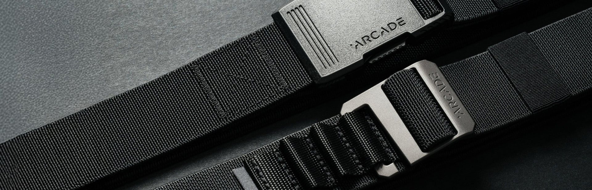 Close-up of two black Arcade brand belts with textured straps and metal buckles, placed parallel on a dark surface.
