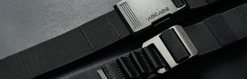 Close-up of two black Arcade brand belts with textured straps and metal buckles, placed parallel on a dark surface.
