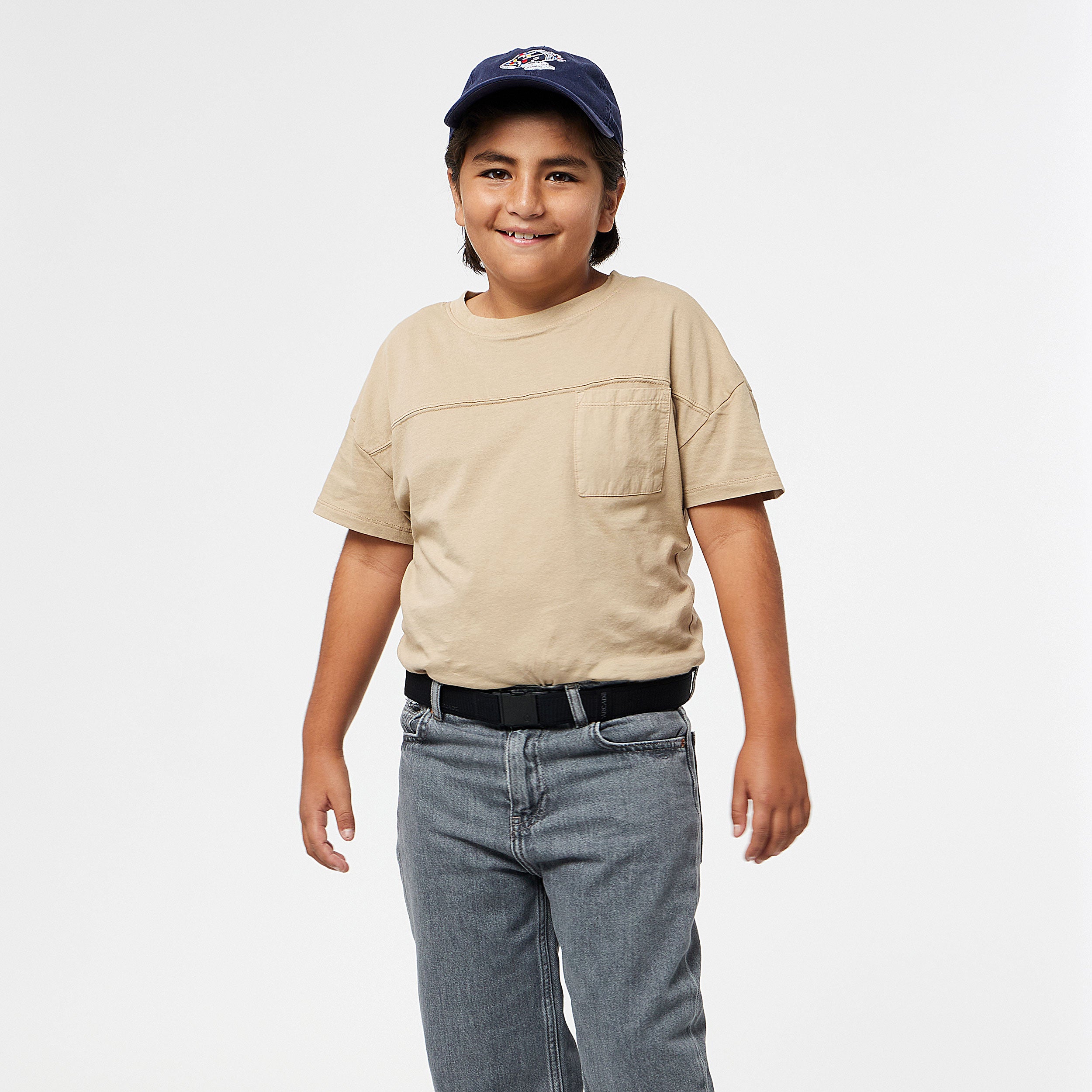 A smiling child with long dark hair wears a navy baseball cap, beige T-shirt, gray jeans, and a black belt from the machine-washable Arcade Belts A2 Youth Kit, standing against a plain white background.