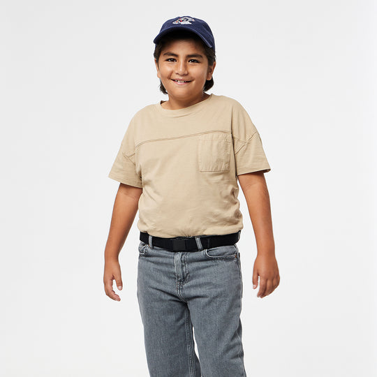 A smiling child with long dark hair wears a navy baseball cap, beige T-shirt, gray jeans, and a black belt from the machine-washable Arcade Belts A2 Youth Kit, standing against a plain white background.