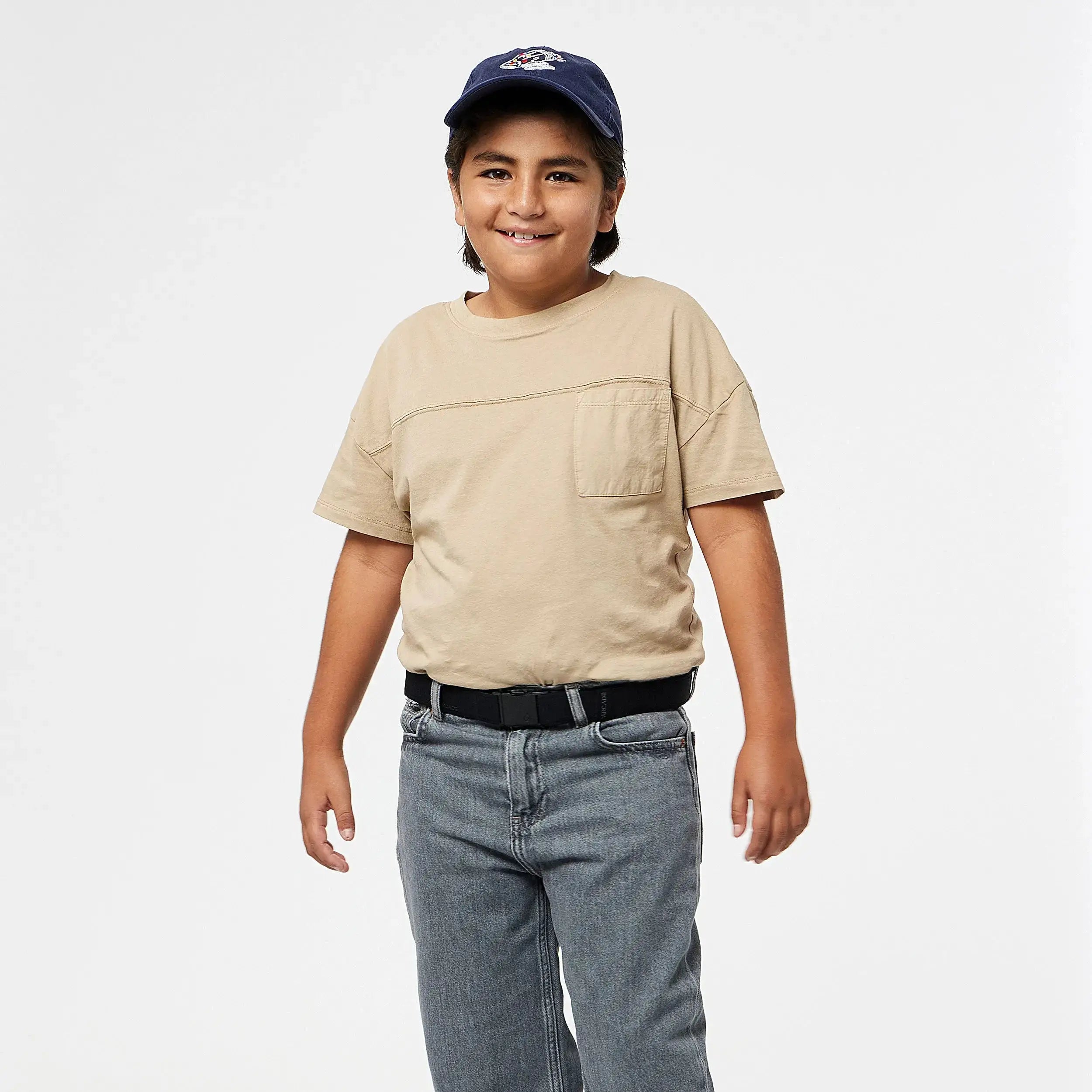A young boy smiles in the A2 Youth Kit from Arcade Belts, featuring a beige tee, blue jeans, black belt, and navy cap—his comfy outfit is machine-washable and shown against a plain white background.