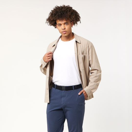 A young man with curly hair models the Arcade Belts Momentum Kit—a beige performance stretch jacket over a white tee and blue pants—standing against a plain light background, one hand holding his jacket lapel.