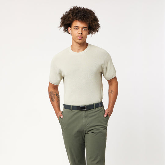 A person with curly hair wears the Arcade Belts Lifestyle Kit: a light beige short-sleeve shirt and versatile olive green pants, posing with hands in pockets against a plain white background.