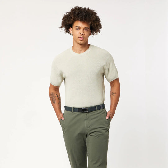 A person with curly hair wears the Arcade Belts Lifestyle Kit: a light beige short-sleeve shirt and versatile olive green pants, posing with hands in pockets against a plain white background.