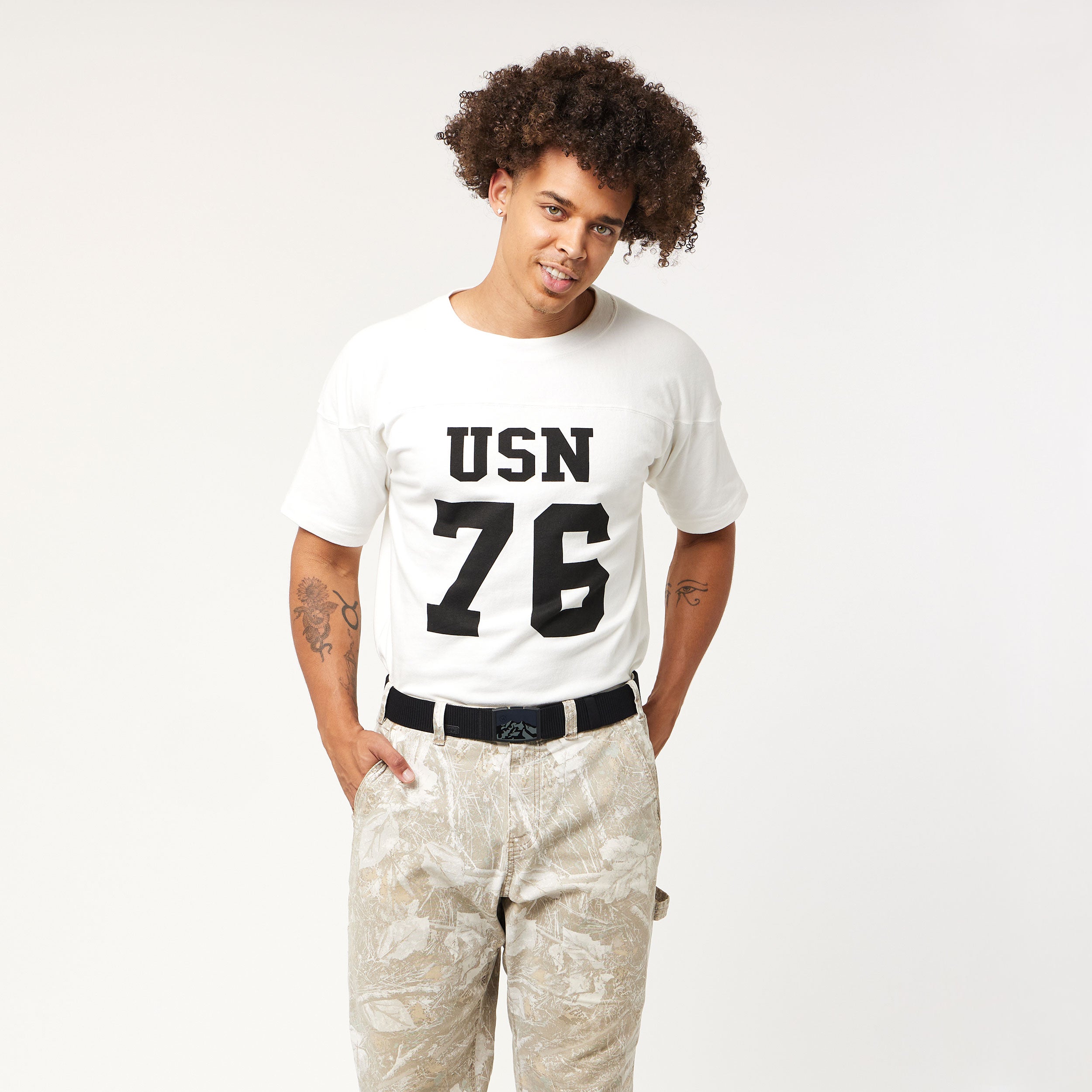 A man with curly hair wears the Arcade Belts Woody Kit—featuring a white t-shirt with USN 76 in bold black letters, light patterned pants, and the iconic belt—standing against a white background, smiling with hands in his pockets.
