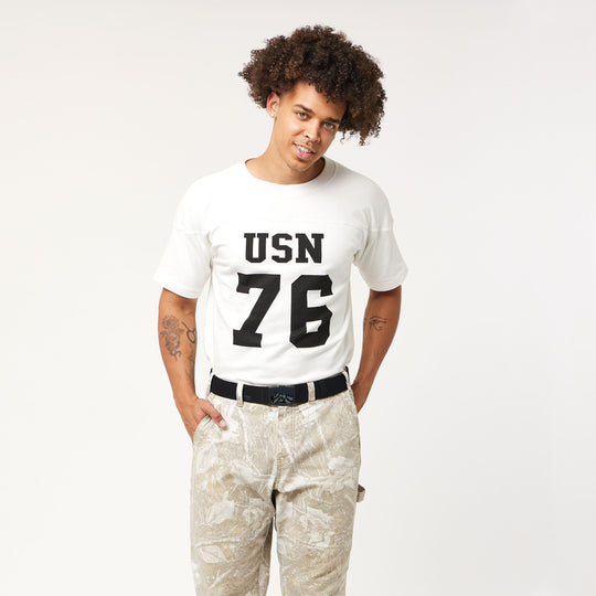 A man with curly hair wears the Arcade Belts Woody Kit—featuring a white t-shirt with USN 76 in bold black letters, light patterned pants, and the iconic belt—standing against a white background, smiling with hands in his pockets.