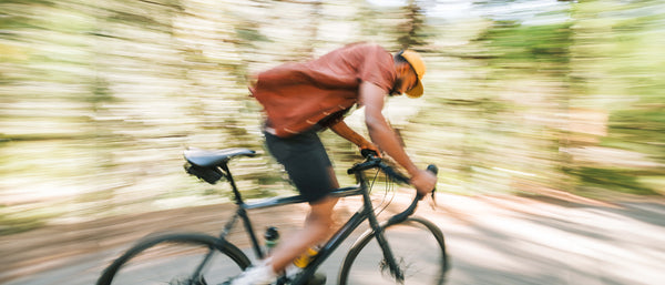A person wearing a brown shirt and yellow cap is riding a bicycle quickly through a forest, sporting an Arcade Belt with a durable plastic buckle—the blurred background highlights the thrill of adventure in motion.