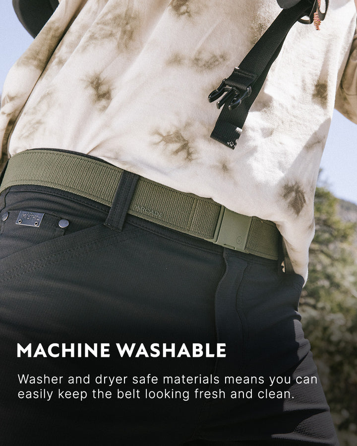 A person wearing black pants, an olive green Elastic Stretch Belt, and a beige tie-dye shirt stands outdoors. Text at the bottom reads, Machine Washable: Washer and dryer safe materials mean you can easily keep the belt looking fresh and clean.
