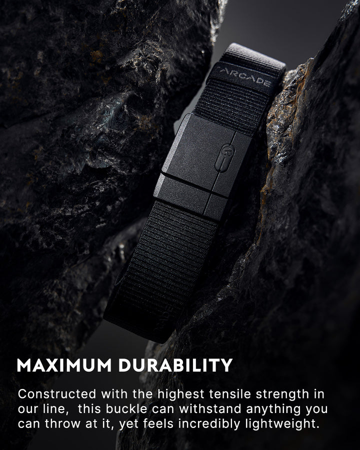 A black Arcade belt with a durable buckle is positioned upright between two dark rocks. The accompanying text highlights the belt’s maximum durability and lightweight design.