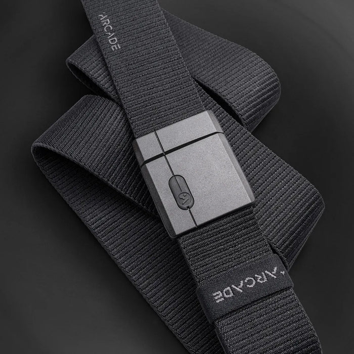 A black fabric belt with a rectangular, matte black buckle is displayed on a dark surface. The belt has the word ARCADE printed on one end in white, uppercase letters.