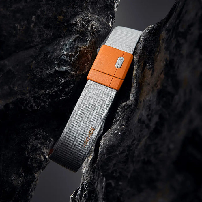A gray fabric belt with an orange buckle, featuring the word ARCADE, is positioned vertically between two dark, textured rocks, creating a dramatic contrast.