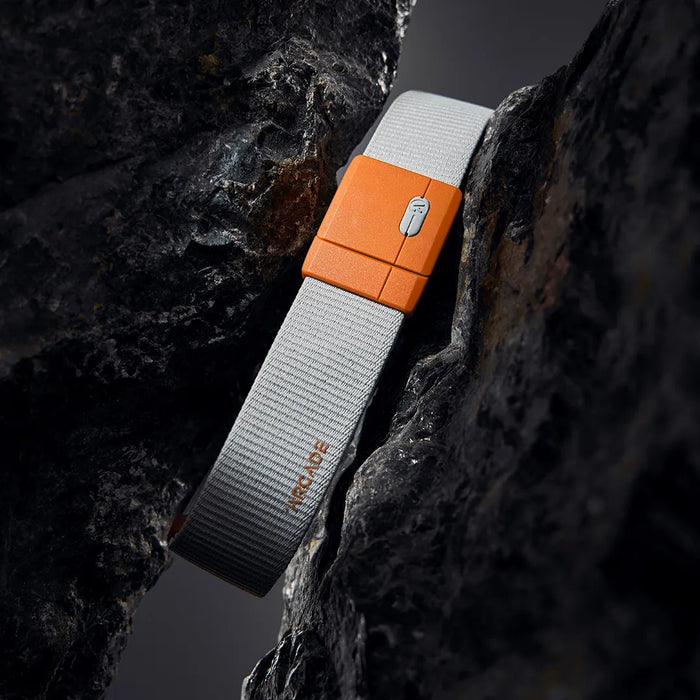 A gray fabric belt with an orange plastic buckle is positioned between two dark, textured rocks. The word ARCADE is visible on the belt near the buckle.
