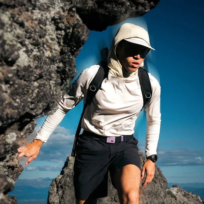 A person wearing a light sun hat, sunglasses, a long-sleeve shirt, and shorts climbs between rocks on a mountain, with a blue sky and distant landscape in the background.