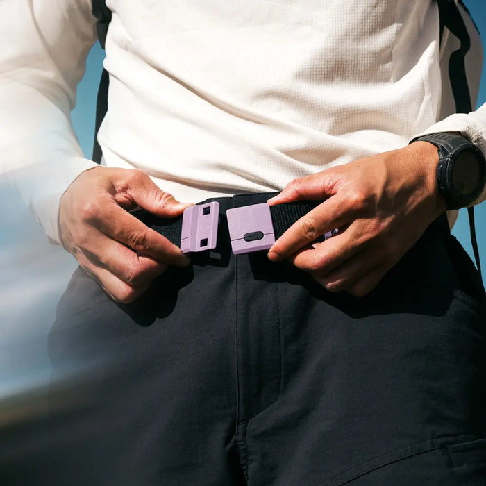 A person wearing a white long-sleeve shirt and black pants fastens a purple buckle on a black belt, with a watch visible on their left wrist.