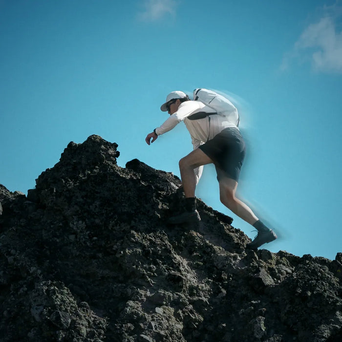 A person wearing a light shirt, shorts, hat, and backpack climbs up a steep, rocky hill against a clear blue sky, with a motion blur effect emphasizing movement.