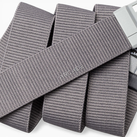 A close-up of a gray, textured Aero Mag belt by Arcade Belts with a durable metal buckle. The word “ARCADE” is embossed on the belt, which is arranged in overlapping folds.