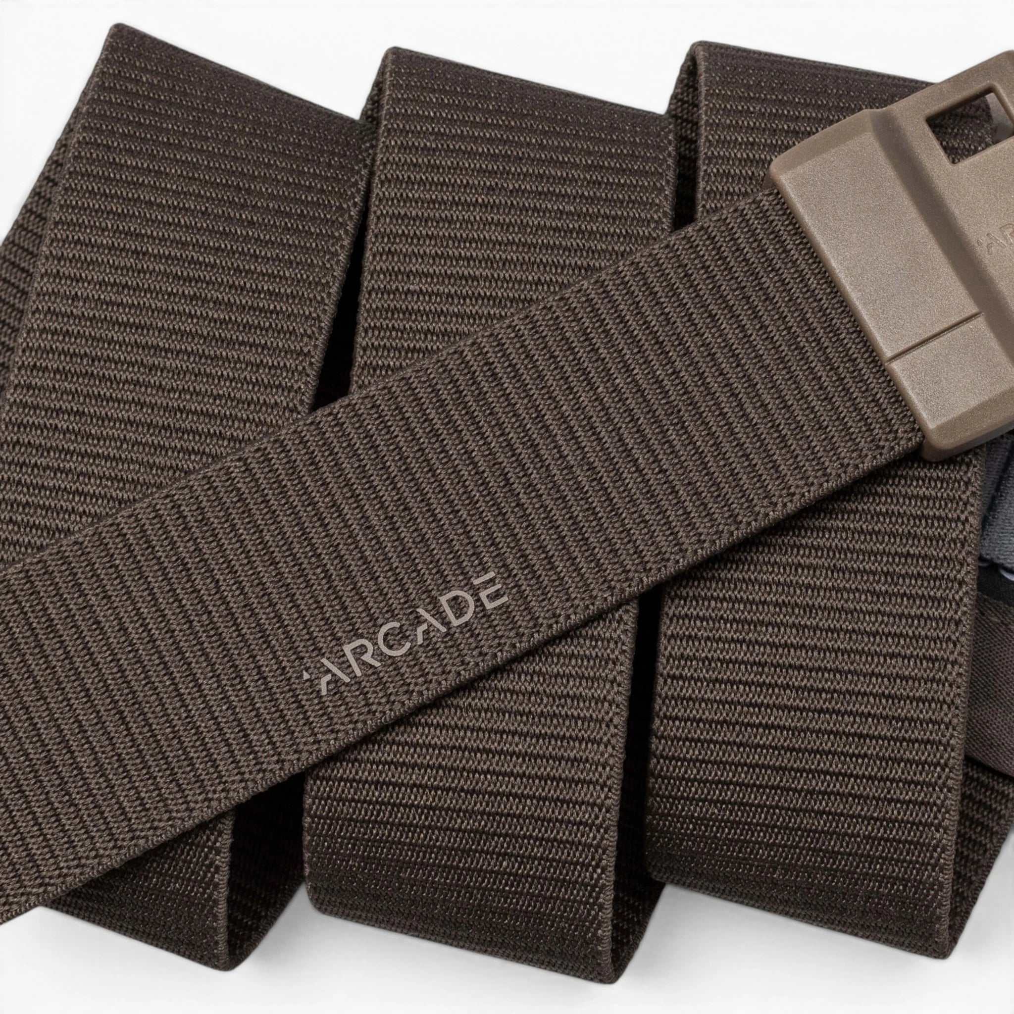 A close-up of the Arcade Belts Aero Mag, a brown woven fabric belt featuring a durable buckle and a rectangular plastic design. The word "ARCADE" is printed on the belt, which is coiled in the background.