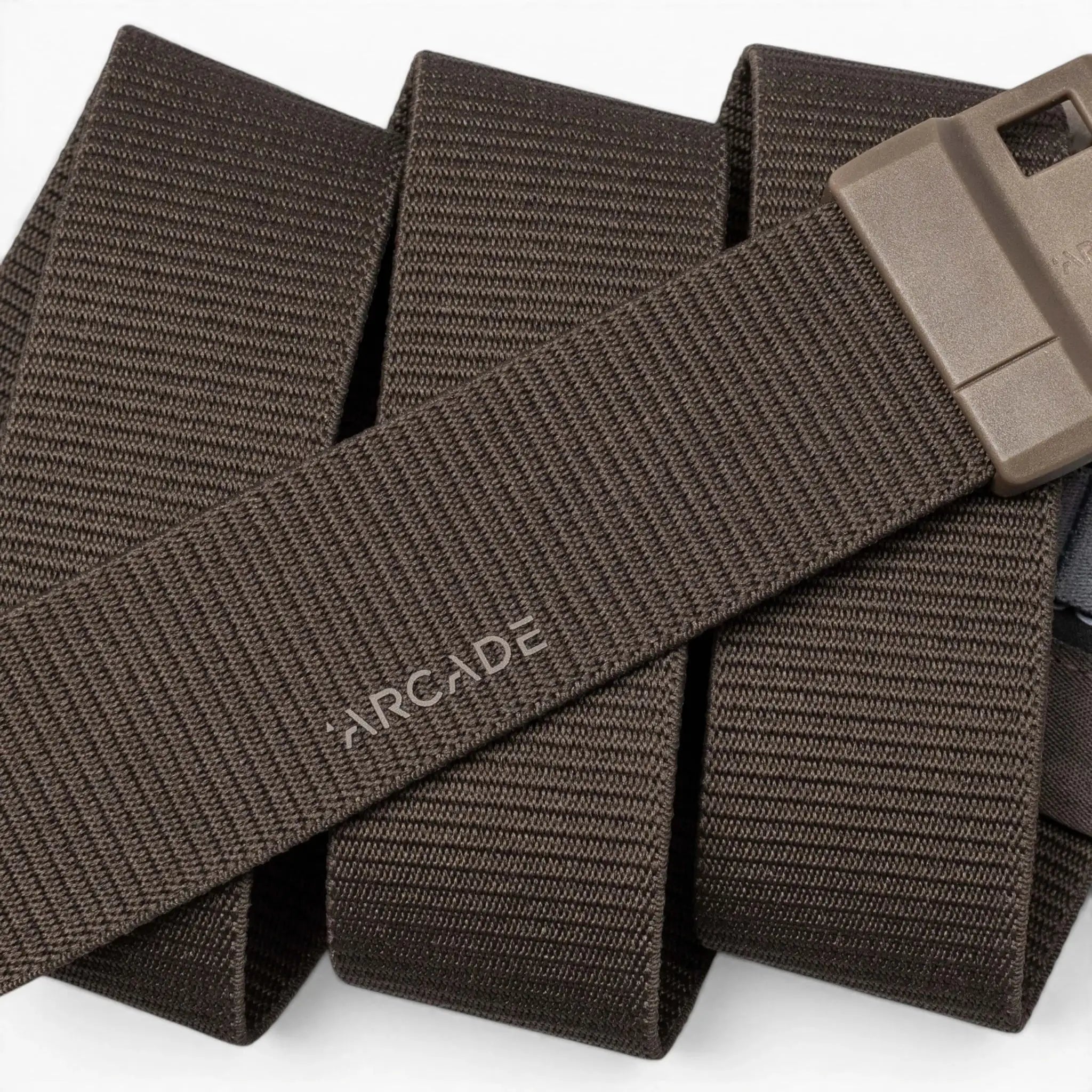 A close-up of the Arcade Belts Aero Mag, a brown woven fabric belt featuring a durable buckle and a rectangular plastic design. The word "ARCADE" is printed on the belt, which is coiled in the background.
