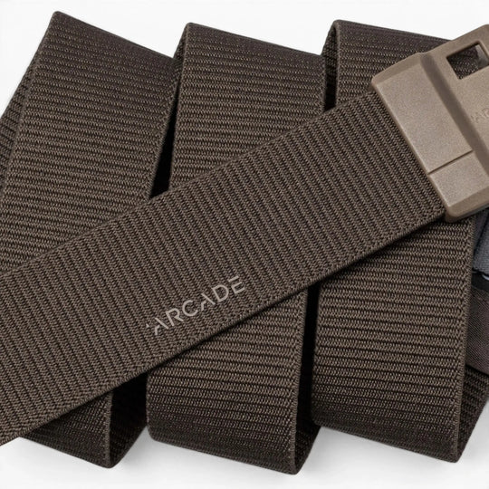 A close-up of the Arcade Belts Aero Mag, a brown woven fabric belt featuring a durable buckle and a rectangular plastic design. The word "ARCADE" is printed on the belt, which is coiled in the background.